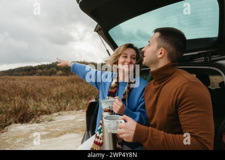 Heureux jeune femme et homme assis dans le coffre ouvert d'une voiture tout en voyageant en automne, concept de voyage routier Banque D'Images