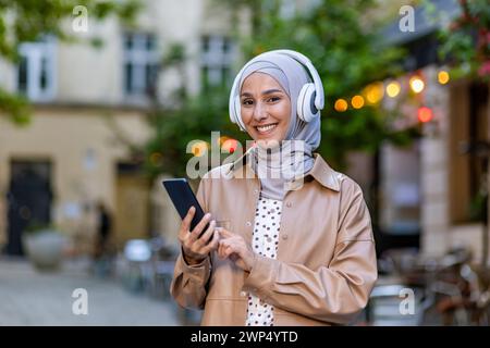 Charmante femme musulmane portant des écouteurs sur le hijab et utilisant le téléphone portable pendant la promenade en ville. Belle dame positive appréciant écouter de la musique préférée via des appareils électroniques sans fil. Banque D'Images