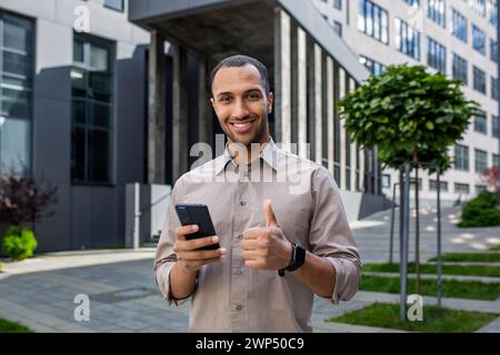 Homme musulman optimiste montrant un geste du pouce vers le haut tout en utilisant un smartphone et en souriant à la caméra à l'extérieur. Fier gars recevant une notification agréable sur les cours des actions en hausse tout en se tenant au centre-ville. Banque D'Images