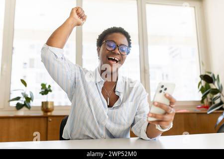 Femme afro-américaine ravie regardant le téléphone et célébrant le succès assis au bureau avec smartphone. Jeune employée excitée par les bonnes nouvelles dans l'email ou le message, promotion d'emploi, remboursement d'argent Banque D'Images