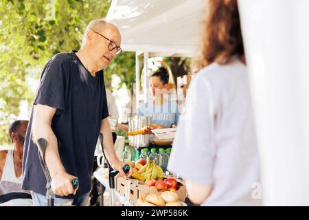 Les efforts caritatifs à la collecte de nourriture comprennent l'aide à un vieil homme caucasien défavorisé sur des béquilles avec le soutien de bénévoles de diverses ethnies. Un homme handicapé reçoit une aide humanitaire. Banque D'Images