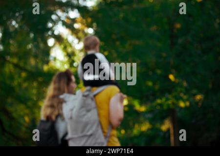 Photo floue avec une jeune famille - maman, papa, enfant de 1-2 ans marchant dans un parc de printemps. Heureux parents d'un tout-petit. Papa portant un bébé sur un dos. Banque D'Images