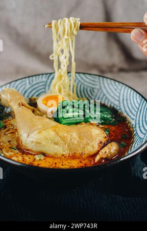 Un bol de soupe avec des nouilles et du poulet dedans. La soupe est orange et verte. Une personne mange la soupe avec des baguettes Banque D'Images