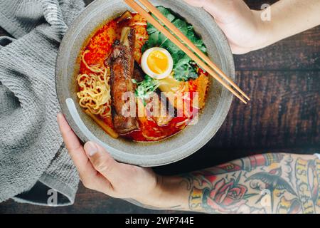 Une personne tient un bol de ramen avec des baguettes. Le bol est rempli de nouilles, de viande et d'œufs. La personne semble apprécier son repas Banque D'Images