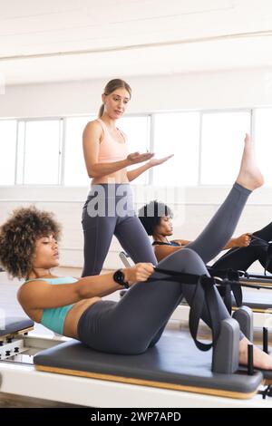 Jeune femme caucasienne enseigne aux femmes biraciales dans un gymnase sur les réformateurs de pilates Banque D'Images