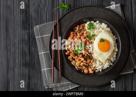 pad krapow gai, pad gaprao, pad kra pao, poulet sauté au basilic avec riz et oeuf frit sur assiette avec baguettes sur table en bois noir, cuisine thaïlandaise, Banque D'Images