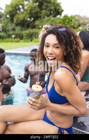 Femme biraciale dans un maillot de bain bleu aime une fête de la piscine, tenant un verre avec un sourire Banque D'Images