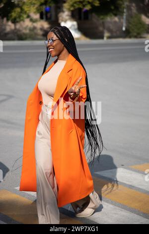 Kazakhstan, Shymkent - 04 septembre 2023 : femme africaine joyeuse . Dame brune aux cheveux longs en manteau orange sourit et se promène à l'extérieur. Banque D'Images