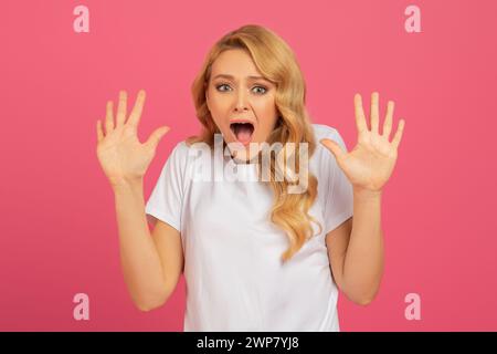 femme blonde lève les mains dans un geste choqué sur fond rose Banque D'Images