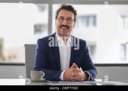Homme d'affaires confiant et joyeux avec une moustache portant un costume bleu assis à un bureau Banque D'Images