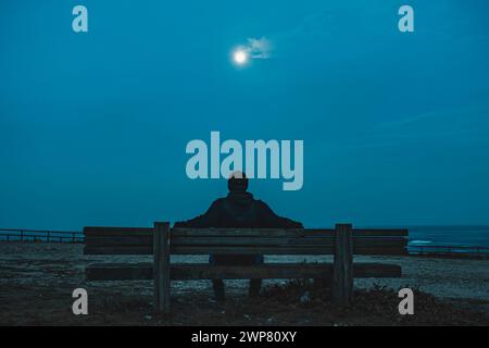 Un homme assis sur un banc regardant l'horizon avec la lune sur le dessus, les tons bleu foncé et l'espace de copie Banque D'Images