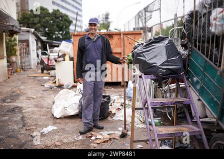 Homme, travail et nettoyage des ordures pour la gestion des déchets en ville avec portrait, balai et bonheur. Personne, visage et sourire par site de benne à ordures pour Banque D'Images