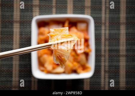 Focus sélectif sur les baguettes en bambou avec une salade de chou kimchi fermenté et un bol de nourriture blanche sur fond brun. Banque D'Images