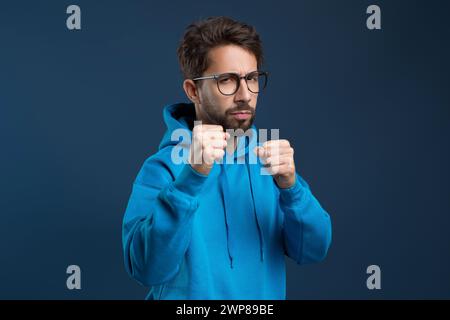 Homme déterminé portant des lunettes et un sweat à capuche levant les poings en posture défensive Banque D'Images