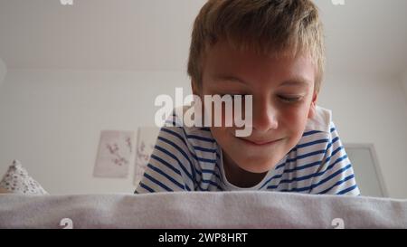 Mignon petit garçon caucasien de 10 ans se trouve à la maison sur le lit dans la chambre et sourit slyly, plissant les yeux et fermant les yeux. Je m'attends à une surprise Banque D'Images