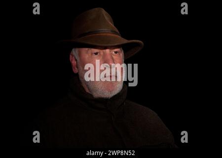Portrait d'un vieux chasseur réfléchi avec une barbe qui respire le calme et son visage marqué par l'expérience, sur un fond noir. Banque D'Images