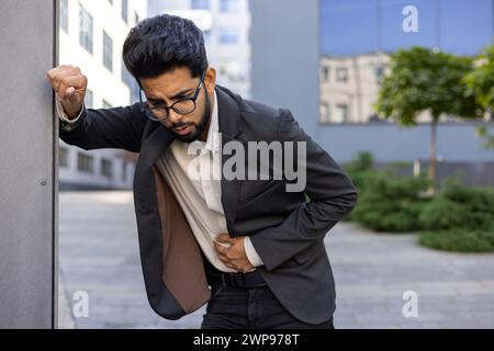 Un jeune homme indien professionnel vêtu d'une tenue d'affaires chic et décontractée, debout à l'extérieur, exprimant l'inconfort et la douleur en serrant son ventre. Cette image capture un moment de détresse potentiel maladie. Banque D'Images