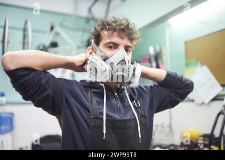 Portrait d'un jeune travailleur latin ajustant un masque de protection respiratoire, avant de commencer un travail de peinture dans son atelier. De vraies personnes au travail. Banque D'Images