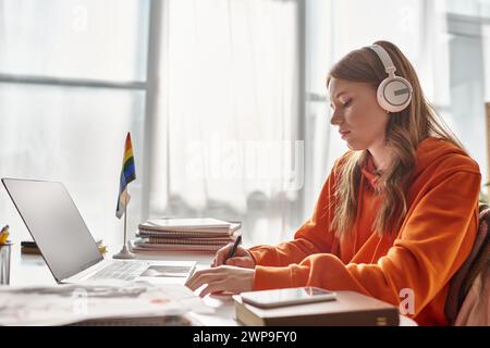 jeune adolescente concentrée dans des écouteurs sans fil e-learning à côté du drapeau de fierté et de la papeterie Banque D'Images