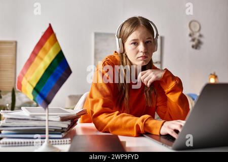 jeune adolescente fatiguée dans des écouteurs sans fil en utilisant son ordinateur portable à côté du drapeau de fierté et de la papeterie Banque D'Images