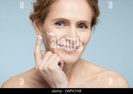 Femme mature positive applique une crème hydratante sur son visage, sur fond bleu. Crème pour le visage hydratant la peau, soin de la peau Banque D'Images