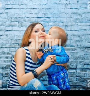 Une jeune belle mère est assise avec son enfant sur le fond d'un mur de briques bleues. Maman embrasse son fils. Banque D'Images