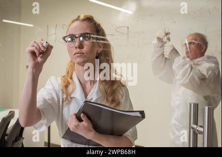 Une femme chimiste écrit une formule sur verre. Un homme caucasien âgé en costume de protection fait des tests. Banque D'Images