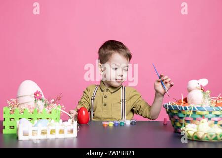 Charmant jeune avec un sourire fabriquant des œufs et des décorations pour les festivités de Pâques, en utilisant des fournitures d'art et de la peinture à l'aquarelle. Petit garçon heureux se sentant excité par l'activité de peinture. Banque D'Images