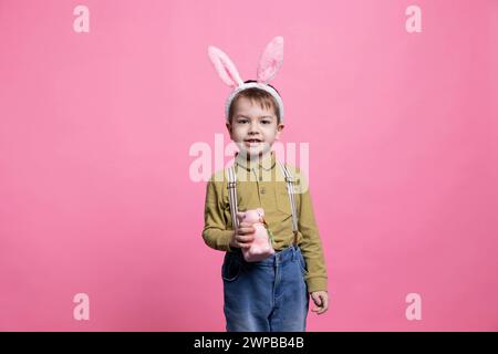 Doux jeune garçon joyeux posant avec un jouet de lapin moelleux sur la caméra, portant des oreilles de lapin et célébrant la fête des vacances de pâques. Petit enfant étant excité et joyeux sur fond rose. Banque D'Images