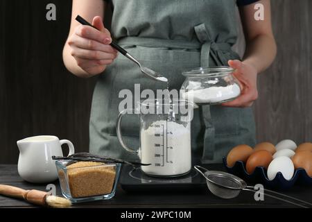 Femme ajoutant de la levure chimique dans la tasse à mesurer à la table en bois noir, gros plan Banque D'Images