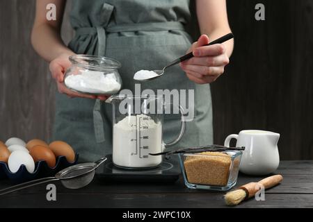 Femme ajoutant de la levure chimique dans la tasse à mesurer à la table en bois noir, gros plan Banque D'Images