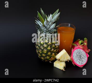 Un verre d'un mélange de jus de fruits fraîchement pressés sur un fond noir, à côté de morceaux d'ananas mûr et d'un demi pitaya. Gros plan. Banque D'Images