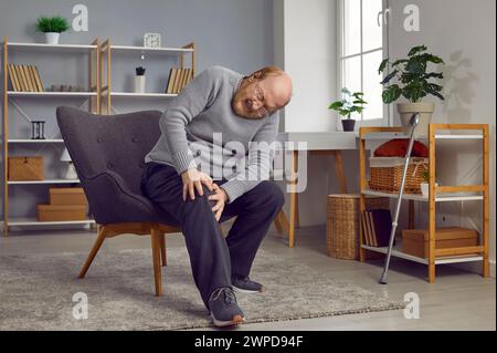 L'homme âgé qui souffre de douleurs au genou est assis sur une chaise et tient son genou Banque D'Images