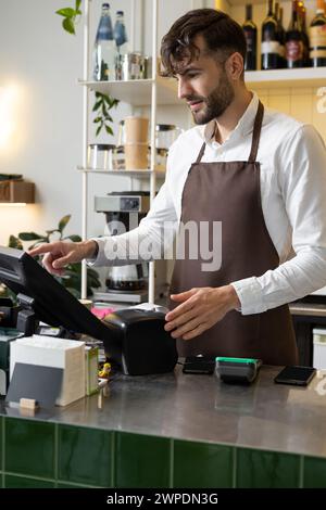 Barista masculin au comptoir en utilisant l'ordinateur de caisse dans le café pour vérifier la commande du client Banque D'Images