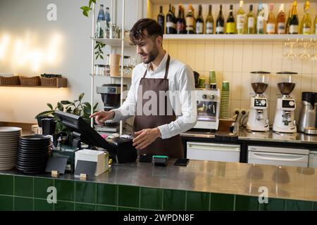 Barista masculin au comptoir en utilisant l'ordinateur de caisse dans le café pour vérifier la commande du client Banque D'Images