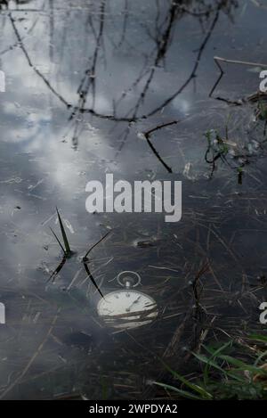 Une vieille montre de poche argentée posée dans une eau peu profonde sur le bord d'un étang, des nuages se reflétant dans l'eau. Banque D'Images