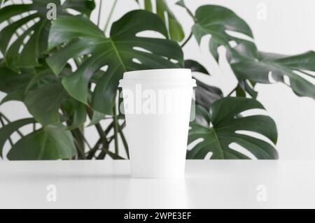 Maquette de tasse de papier de café sur une table blanche avec une plante monstera. Banque D'Images