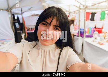 Portrait d'une jeune femme voyageuse latine avec de longs cheveux noirs et des vêtements blancs, en vacances prenant photo selfie avec son téléphone regardant la caméra smilin Banque D'Images