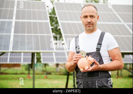 Homme mettant de l'argent dans la tirelire sur fond de panneaux solaires. Homme adulte faisant des investissements dans l'avenir. Homme compétent investissant dans des sources d'énergie alternatives. Banque D'Images