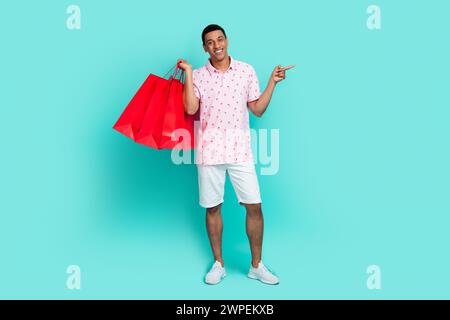 Photo pleine longueur de l'homme de bonne humeur porter polo rose tenir sacs à provisions dirigeant à offrir espace vide isolé sur fond de couleur turquoise Banque D'Images