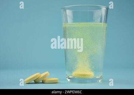 Photographie de couleur orange de vitamine C soluble dans l'eau se dissolvant dans un verre d'eau avec des bulles et des tas de pilules ou de comprimés sur fond bleu Banque D'Images