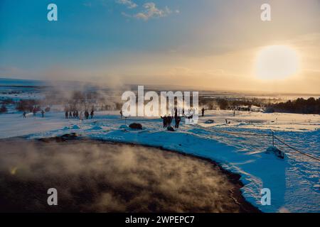 Paysage hivernal avec des sources géothermiques, des touristes et un soleil couchant projetant une lueur chaude sur la scène enneigée. Lieu : Strokkur Geyser, Islande. Banque D'Images