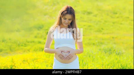 Heureuse jeune femme enceinte fait signe le cœur sur son ventre avec ses mains dans le parc d'été Banque D'Images