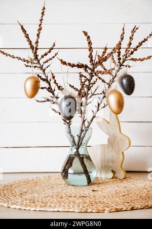 Branches d'arbre dans un vase avec des oeufs de Pâques de couleur noire et bronze suspendus sur une ficelle, fond de planche de bois rustique blanc. Décoration de Pâques minimale à la maison. Banque D'Images