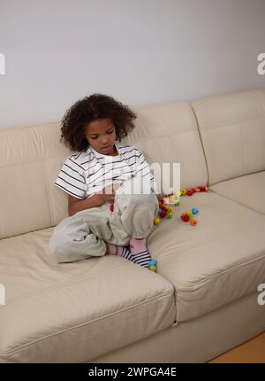 Une jeune fille métisse concentrée sur la couture d'une poupée en tissu sur un canapé crème, entourée de perles colorées. jouer et apprendre, pour les thèmes de l'enfant devel Banque D'Images