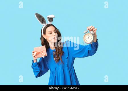 Jeune femme dans le bandeau d'oreilles de lapin de Pâques avec boîte-cadeau et réveil sur fond bleu Banque D'Images