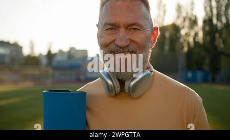 Vieil homme caucasien tenant tapis sportif activité physique pensionné style de vie regardant autour de souriant écouteurs sport stade ville en dehors du fitness Banque D'Images