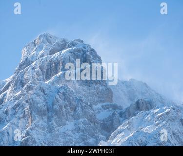 Sommet ensoleillé enneigé dans la vallée de Krnica, Alpes juliennes par une journée d'hiver claire. La brume neigeuse soufflée du sommet par le vent. Banque D'Images