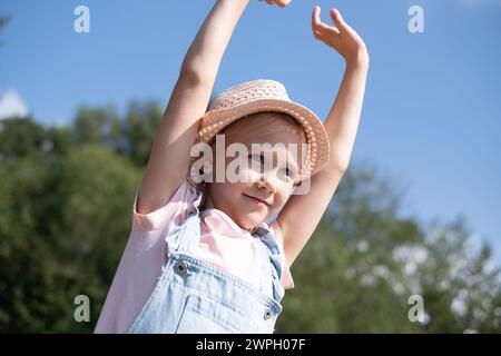 Une joyeuse petite fille portant un chapeau de paille et une salopette en denim étire ses bras contre un ciel bleu, rayonnant de bonheur et de liberté. lié à summ Banque D'Images