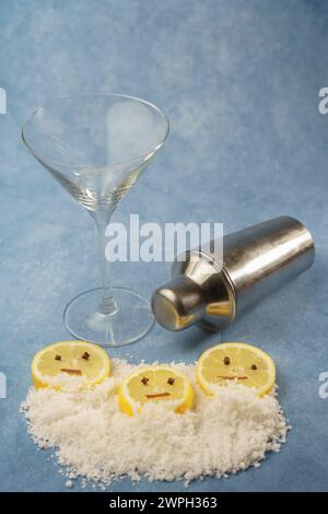 tranches de citron avec clous de girofle et cannelle formant un visage souriant sur un tas de sel à côté d'un shaker à cocktail et un verre de cristal avec fond bleu Banque D'Images